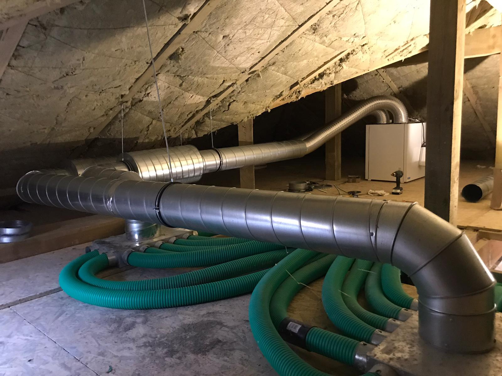 Instalacja rekuperacji w surowym poddaszu: widoczne metalowe rury wentylacyjne podwieszone pod skośnym dachem z izolacją, zielone elastyczne przewody wentylacyjne oraz centrala rekuperacyjna...