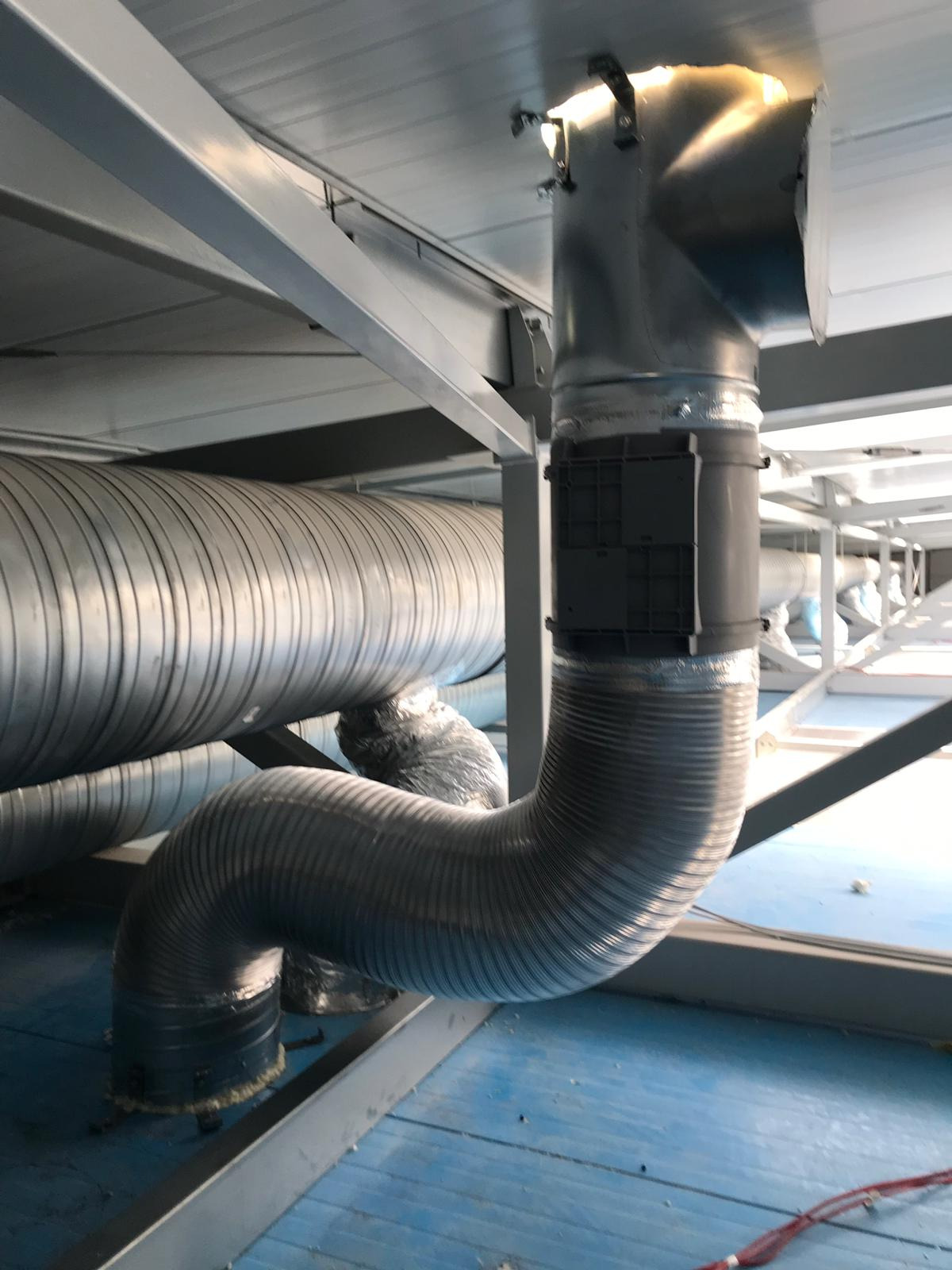 Srebrne, elastyczne kanały wentylacyjne pod sufitem, z widocznymi elementami mocującymi i izolacją, w przestrzeni technicznej z niebieską podłogą.