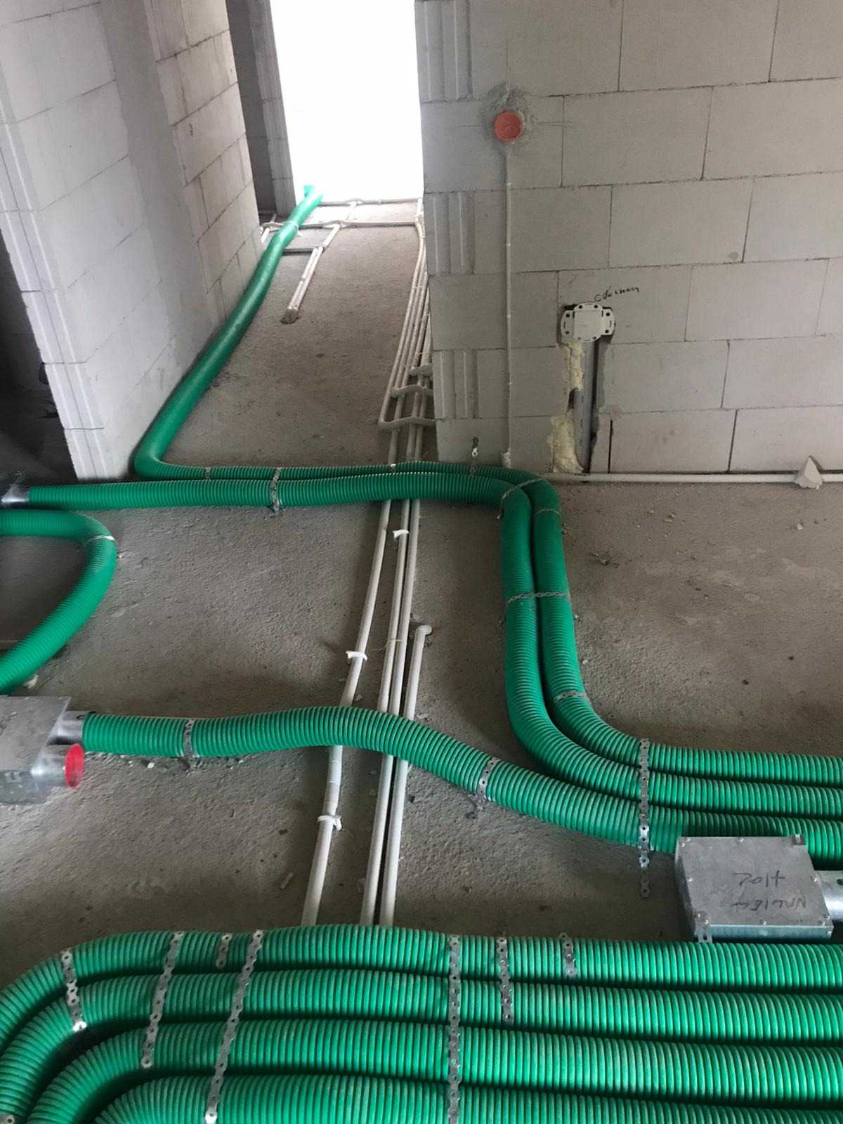 Wnętrze w stanie surowym z widocznymi, zielonymi rurami wentylacyjnymi, przymocowanymi metalowymi uchwytami do podłoża, oraz białymi rurami instalacyjnymi, prowadzącymi w stronę jasnego otworu...