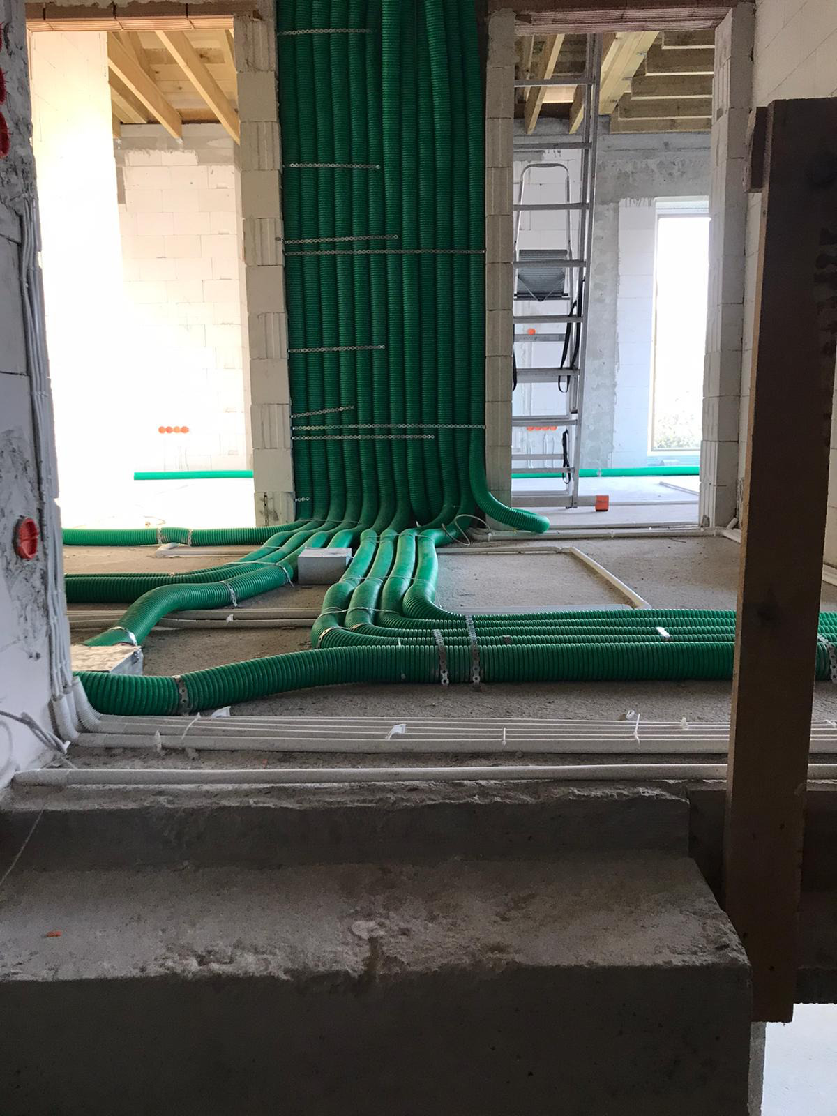Zielone, karbowane rury wentylacyjne prowadzone wzdłuż ściany i podłogi surowego budynku w trakcie montażu, widoczna drabina w tle.