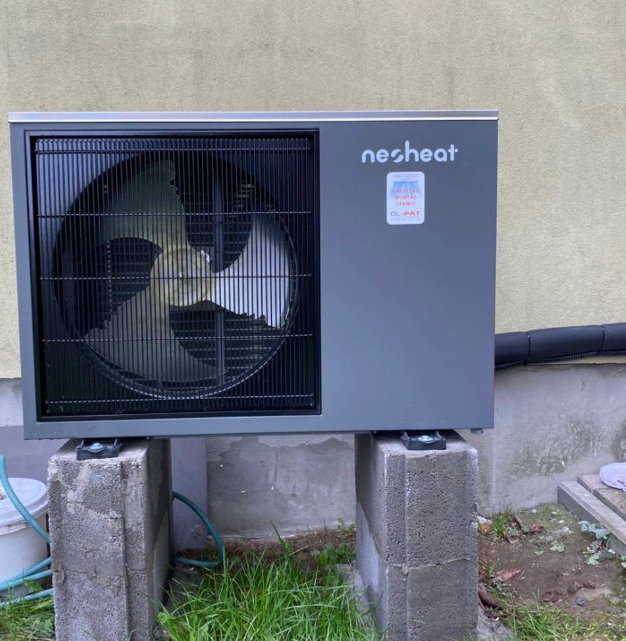 Szara pompa ciepła marki Nesheat zamontowana na betonowych bloczkach, z widocznym wentylatorem za kratką, rurą odpływową i fragmentem trawy.