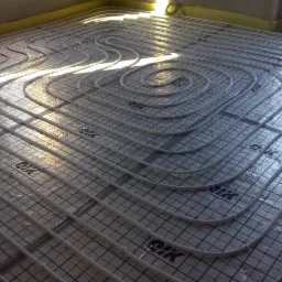 Białe rurki ogrzewania podłogowego ułożone spiralnie na folii z nadrukiem siatki na podłożu w jasnym pomieszczeniu. Widoczne okno i fragment ściany.