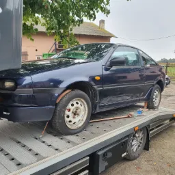Niebieski samochód sportowy z widocznymi śladami użytkowania, zabezpieczony pasami transportowymi na platformie pojazdu pomocy drogowej, z widocznym fragmentem zabudowań w tle.