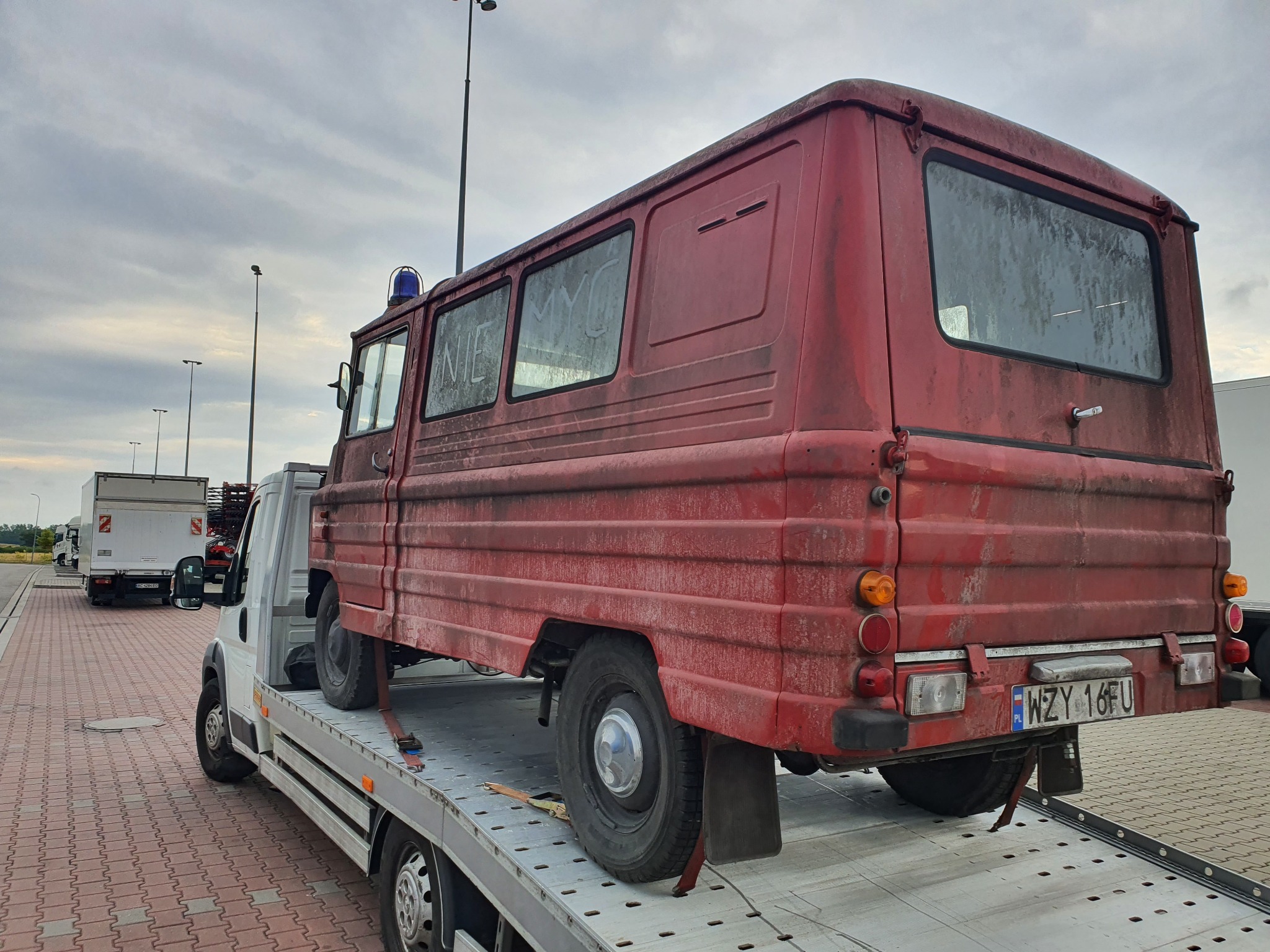 Czerwony, zabytkowy dostawczy samochód z niebieskim kogutem na dachu, transportowany na platformie pojazdu pomocy drogowej. Na oknach widoczne napisy wykonane palcem po brudnej szybie. Tablica...