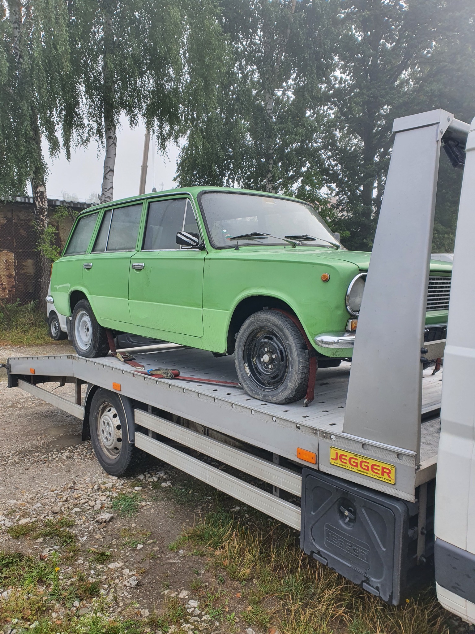 Zielony, klasyczny samochód kombi zabezpieczony pasami na platformie pojazdu transportowego z widocznym logo 'JEGGER' na boku, na tle drzew i szarej kostki brukowej.
