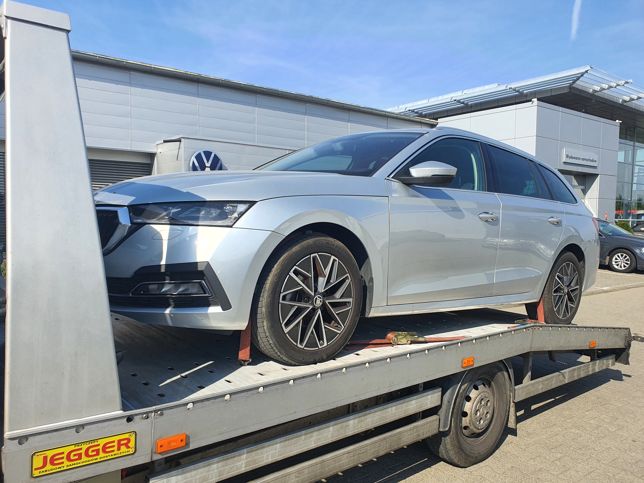 Srebrna Skoda Octavia kombi transportowana na platformie pojazdu z żółtą tabliczką firmy Jegger, na tle budynku salonu samochodowego z logo Volkswagena.