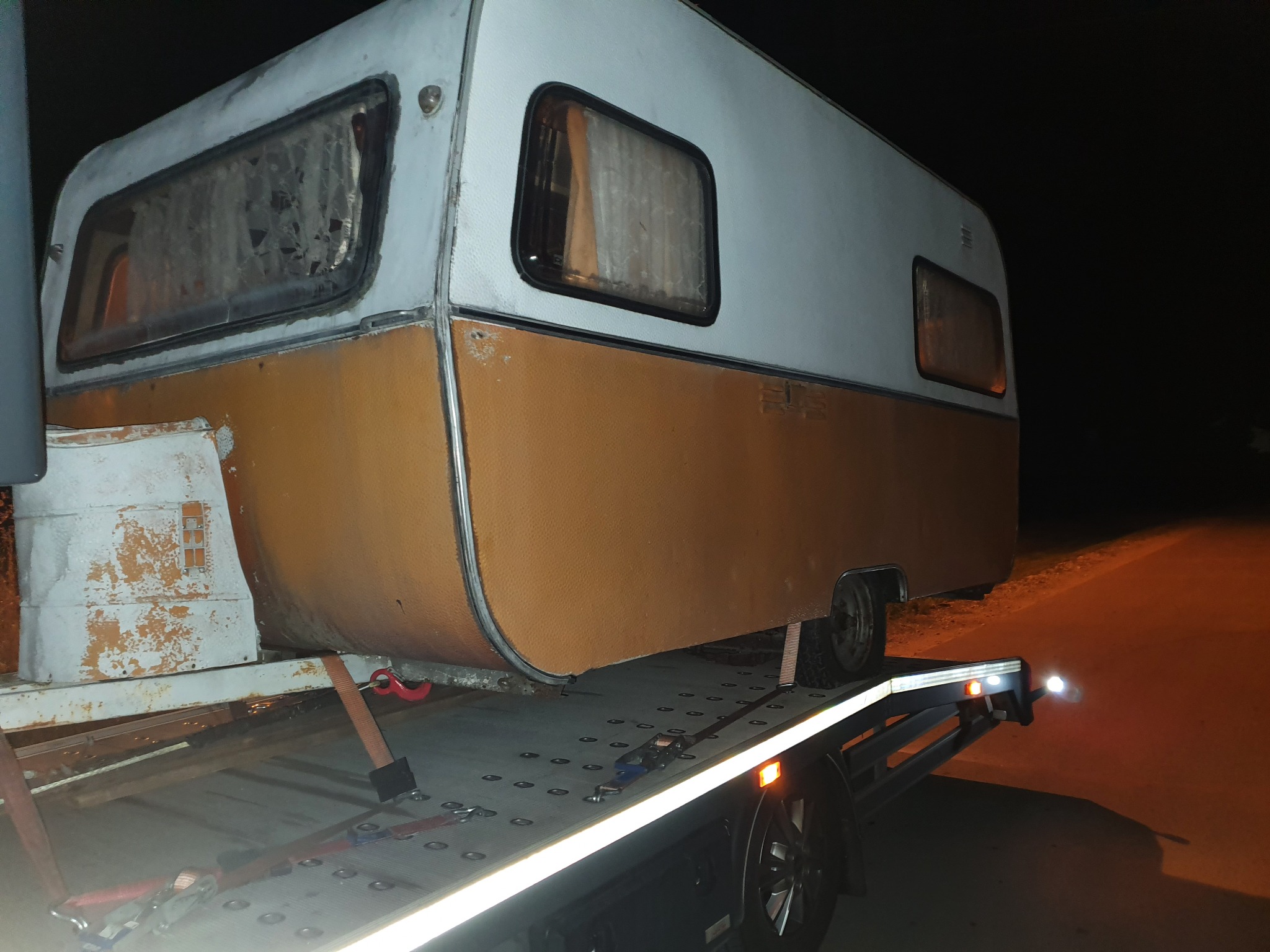 Przyczepa kempingowa z pomarańczowo-białą karoserią, zabezpieczona pasami transportowymi na platformie pojazdu pomocy drogowej, sfotografowana w nocy przy słabym oświetleniu.