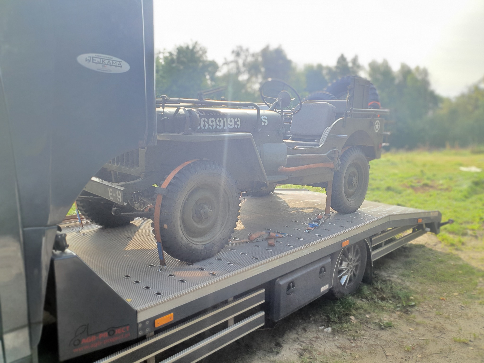 Laweta z wojskowym Jeepem Willys MB zabezpieczonym pasami transportowymi, widoczne detale pojazdu i platformy lawety, tło z zielenią.