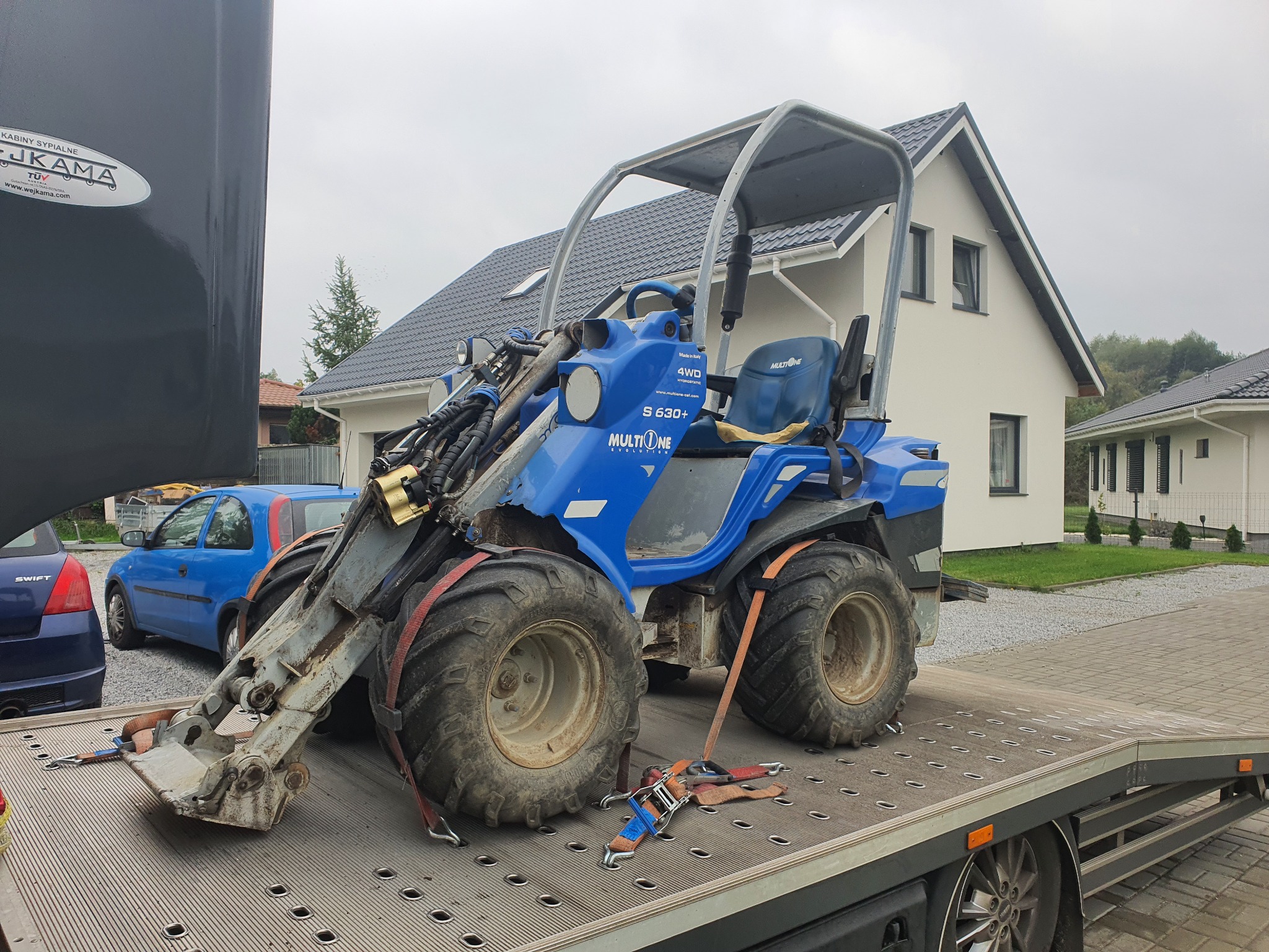 Niebieski miniładowarka Multione S 630+ zabezpieczona pasami transportowymi na platformie pojazdu transportowego, w tle dom jednorodzinny i samochody osobowe.