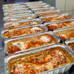 Rząd aluminiowych tacek z gotowym daniem, prawdopodobnie gołąbkami w sosie pomidorowym, posypanych ryżem, ustawionych na metalowym blacie w kuchni.