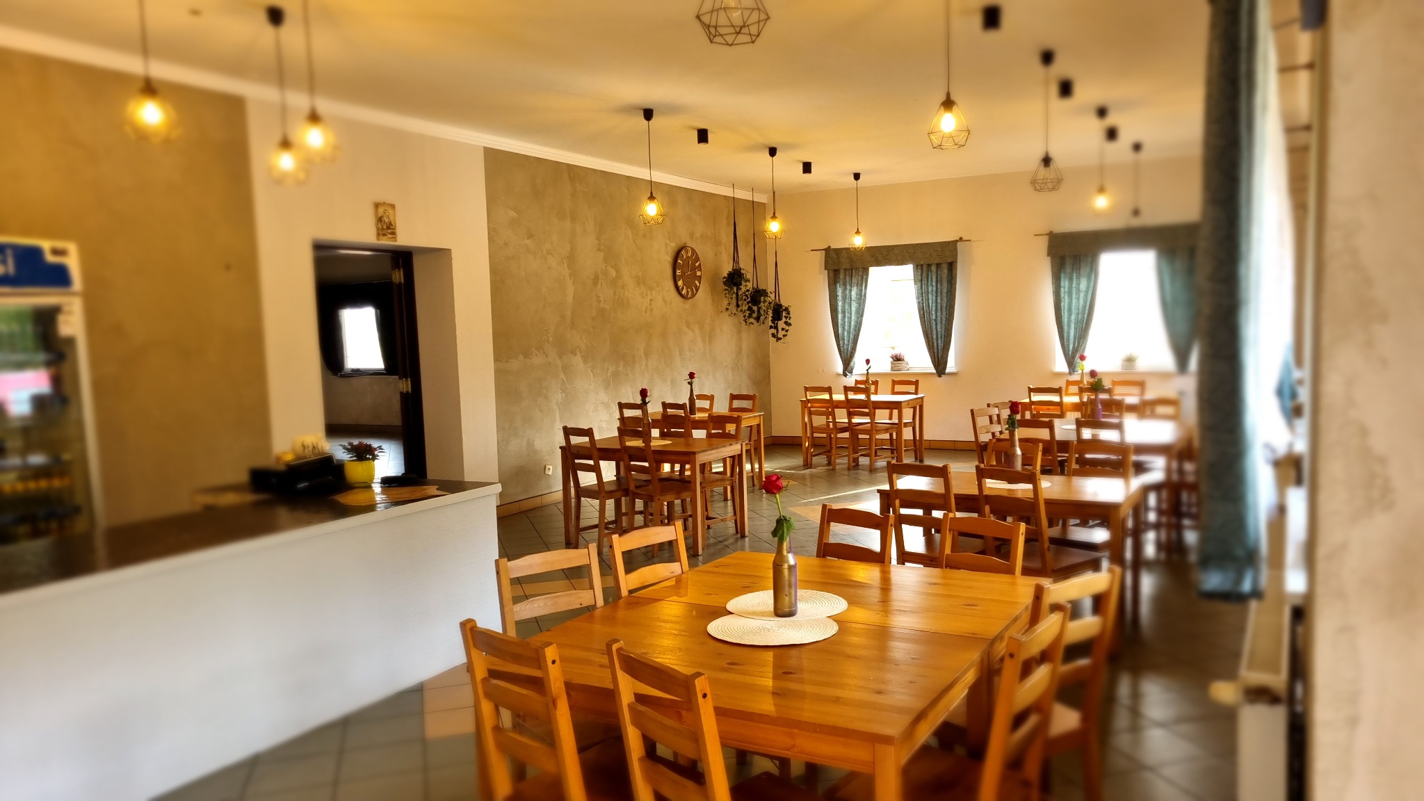 Jasne wnętrze sali restauracyjnej z drewnianymi stołami i krzesłami, dekorowane pojedynczymi czerwonymi różami w wazonach, oświetlone wiszącymi lampami z żarówkami Edisona.