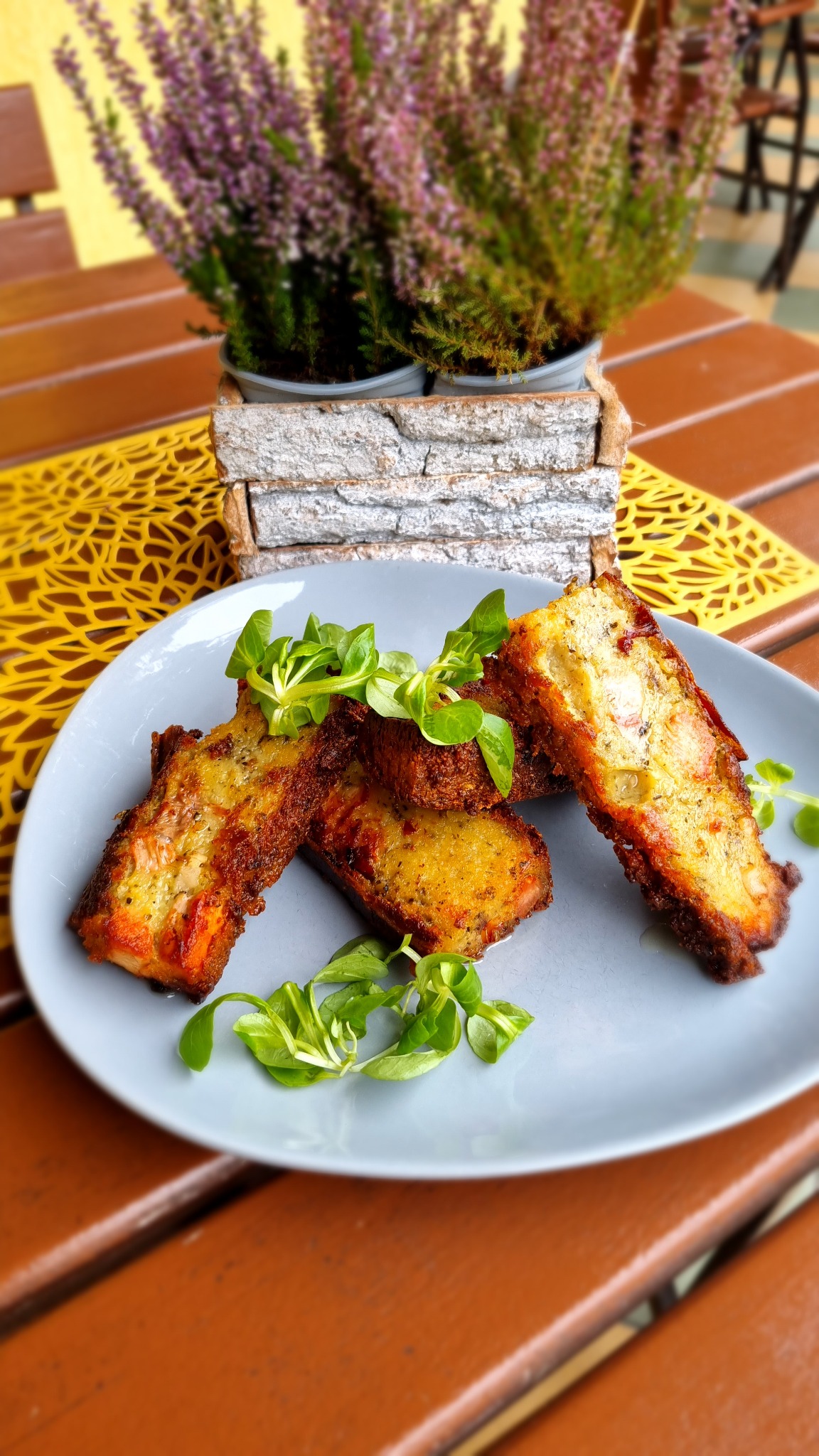 Trzy kromki tostów francuskich, ozdobione świeżymi listkami sałaty, ułożone na szarym talerzu na drewnianym stole z żółtą podkładką, w tle wrzos w drewnianej doniczce.