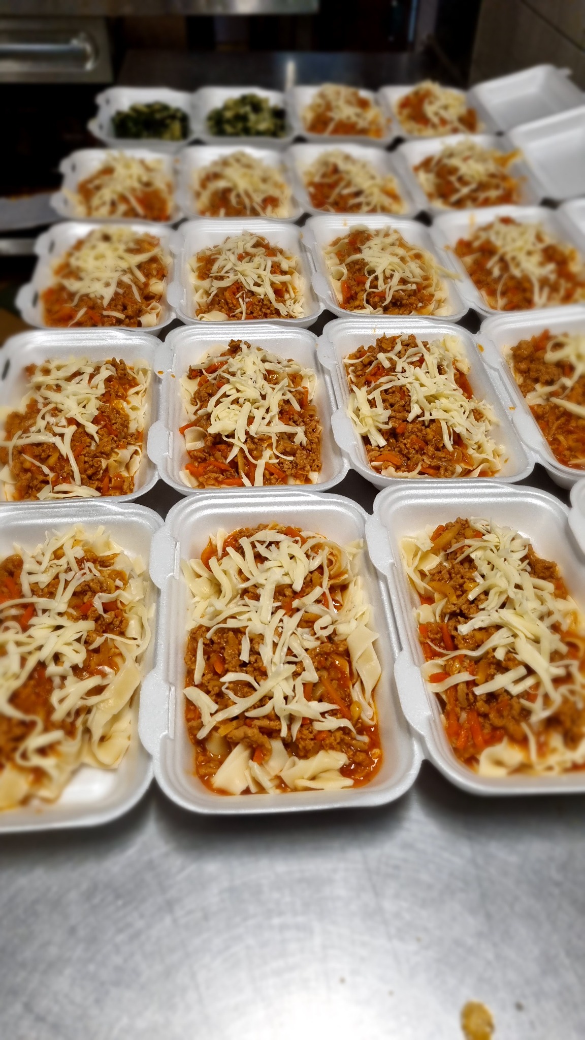 Rzędy styropianowych pojemników z porcjami makaronu z sosem mięsnym i startym serem, przygotowane do transportu.