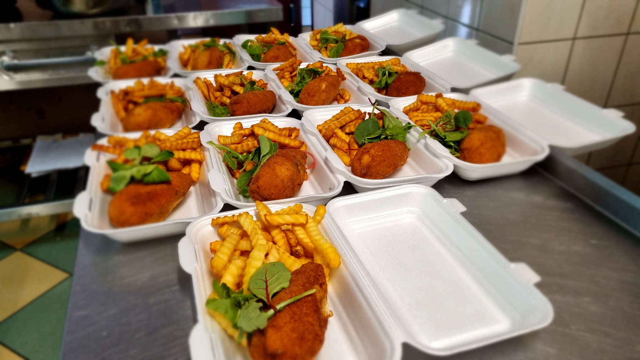 Porcje obiadowe w styropianowych pudełkach: kotlet panierowany, frytki karbowane i zielona sałata, ułożone rzędami na metalowym blacie, gotowe do wydania.