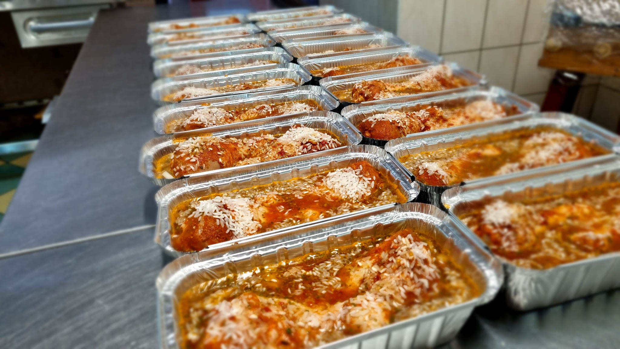 Rząd aluminiowych tacek z gotowym daniem, prawdopodobnie gołąbkami w sosie pomidorowym, posypanych ryżem, ustawionych na metalowym blacie w kuchni.