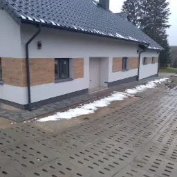 Nowoczesny dom jednorodzinny z białymi ścianami, drewnopodobną okładziną elewacyjną wokół okien, ciemną dachówką i ażurowym podjazdem z betonowych płyt. Na dachu i podjeździe widoczne resztki śniegu.