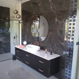 Nowoczesna łazienka z czarnymi płytkami imitującymi marmur, owalnym lustrem i wiszącą szafką z umywalką nablatową oraz złotymi akcentami.