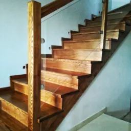 Jasne drewniane schody z balustradą ze szkła mocowanego na metalowych uchwytach, widoczne słoje drewna i płytki podłogowe w odcieniach szarości.