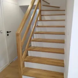 Jasne, drewniane stopnie schodów z białymi podstopnicami i prostą, drewnianą barierką, widoczne z dołu, przy białej ścianie i drzwiach.