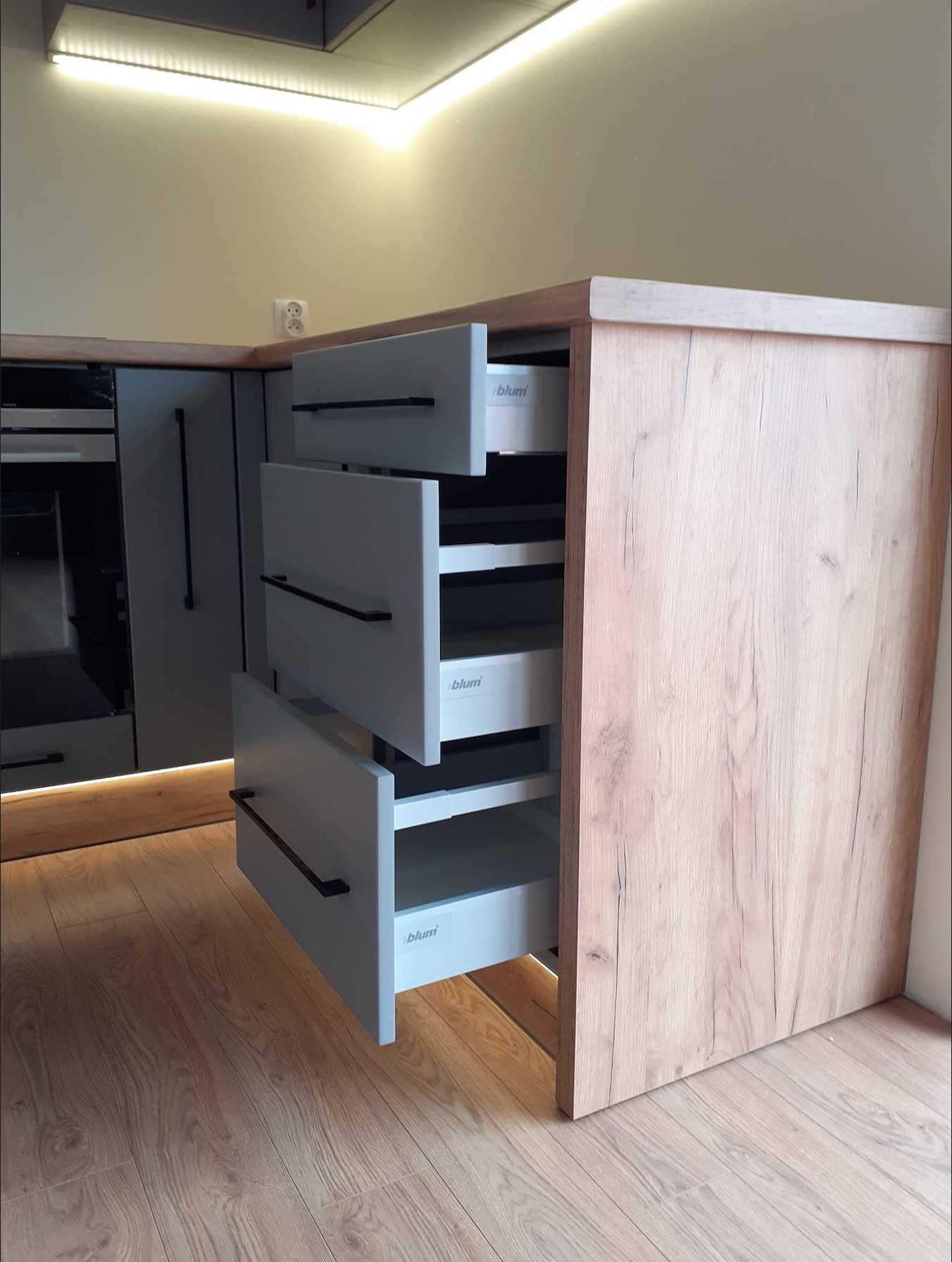 Szare szuflady Blum z czarnymi uchwytami w szafce kuchennej z drewnopodobnym frontem, oświetlenie LED podszafkowe.