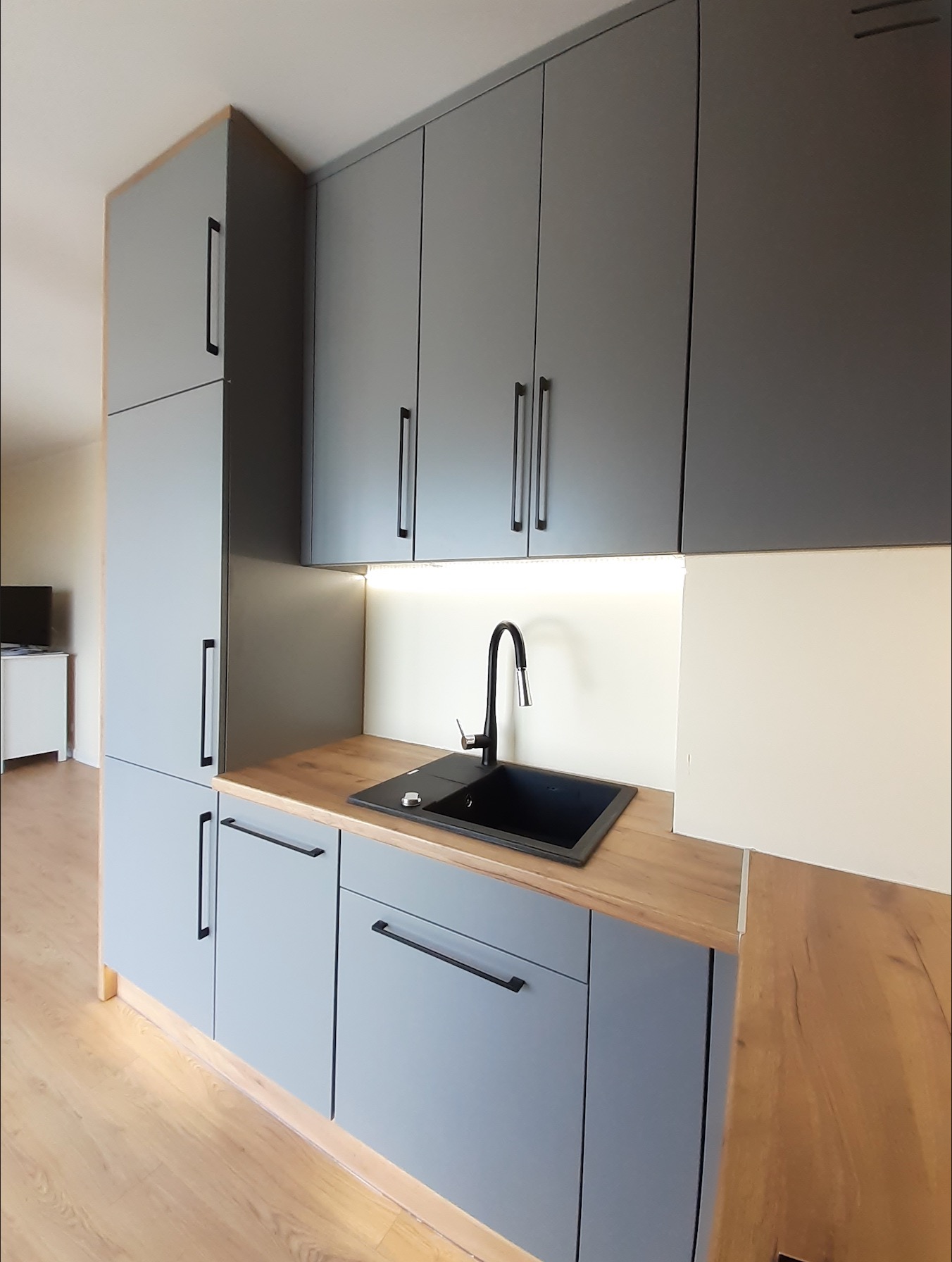 Szara zabudowa kuchenna z drewnianym blatem, czarnym zlewozmywakiem i minimalistyczną armaturą, oświetlenie LED podszafkowe, widok z boku.
