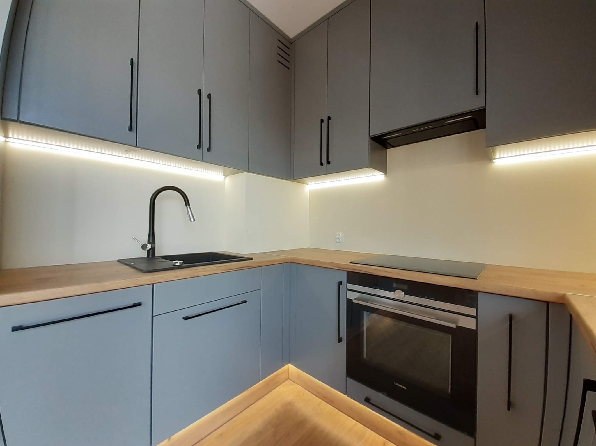 Nowoczesna kuchnia na wymiar z szarymi frontami, drewnianym blatem i oświetleniem LED podszafkowym, widoczny zlew, płyta indukcyjna i piekarnik Siemens.