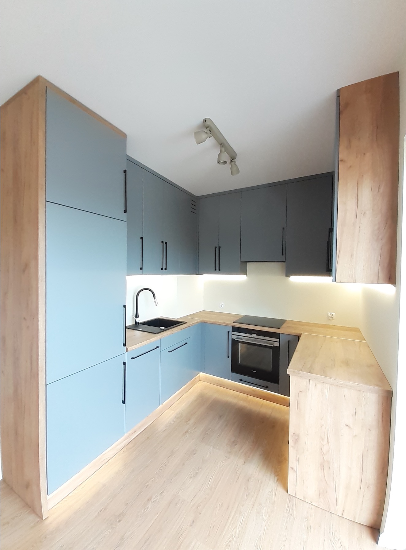 Nowoczesna kuchnia w kształcie litery U z jasnoniebieskimi frontami szafek i drewnopodobnymi bokami, czarny zlew i bateria, oświetlenie LED pod szafkami, płyta indukcyjna.