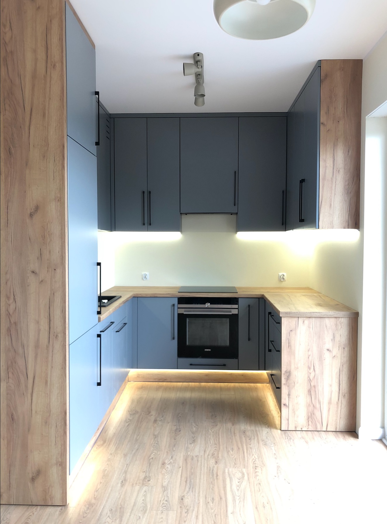 Nowoczesna kuchnia w odcieniach szarości i drewna z podświetleniem LED, minimalistyczne uchwyty, oświetlenie sufitowe i drewnopodobna podłoga tworzą spójną całość.