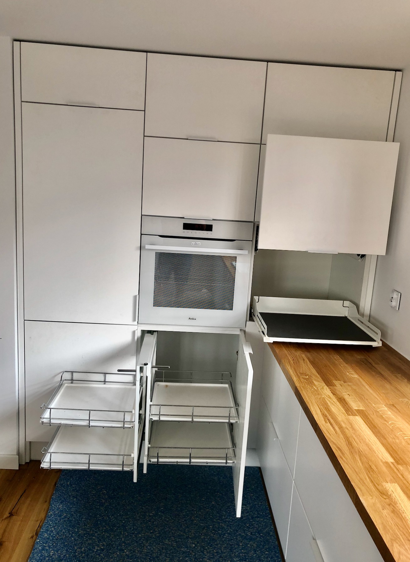 Biała zabudowa kuchenna z piekarnikiem i wysuwanymi koszami cargo, widoczny fragment drewnianego blatu i niebieskiej podłogi.
