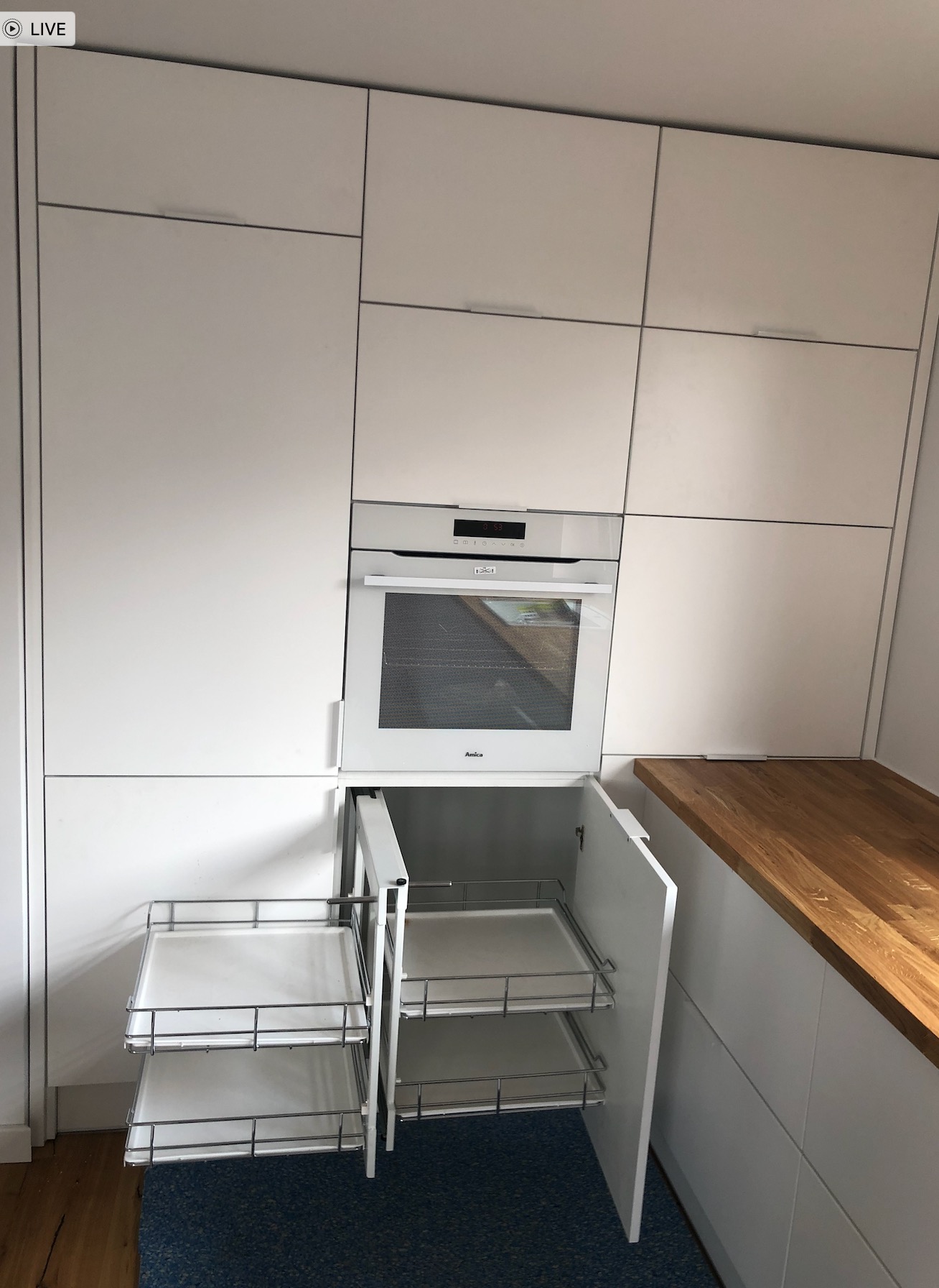 Biała zabudowa kuchenna z piekarnikiem Amica i wysuwanymi koszami cargo w szafce dolnej, widoczny fragment drewnianego blatu.