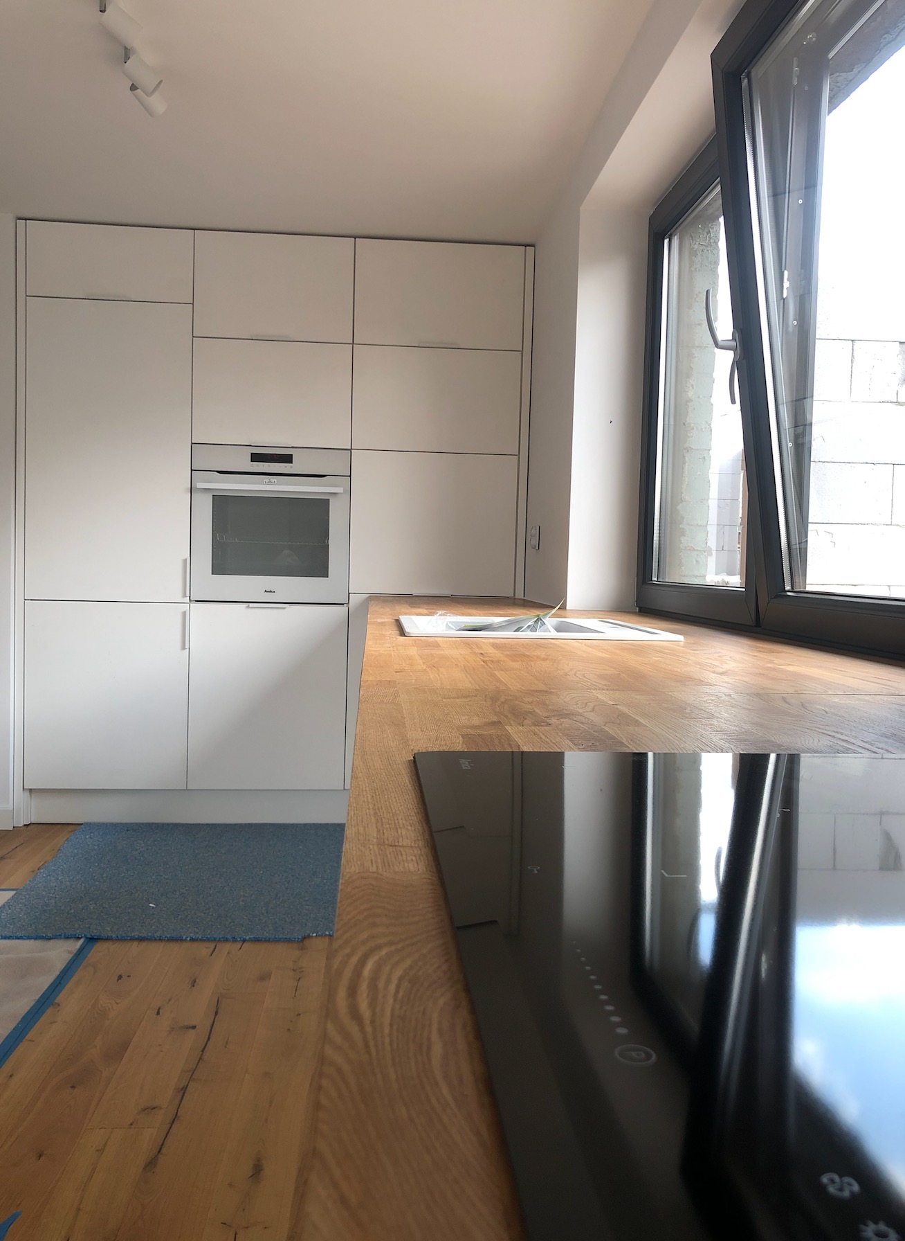 Nowoczesna, jasna kuchnia w Bydgoszczy z białymi frontami szafek, drewnianym blatem, płytą indukcyjną i dużym oknem balkonowym. Na podłodze widoczny fragment niebieskiego dywanika.