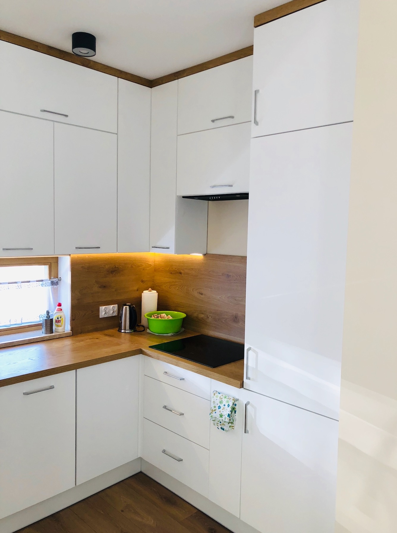 Nowoczesna kuchnia w bieli z drewnianymi akcentami, oświetleniem podszafkowym i zieloną miską na blacie.