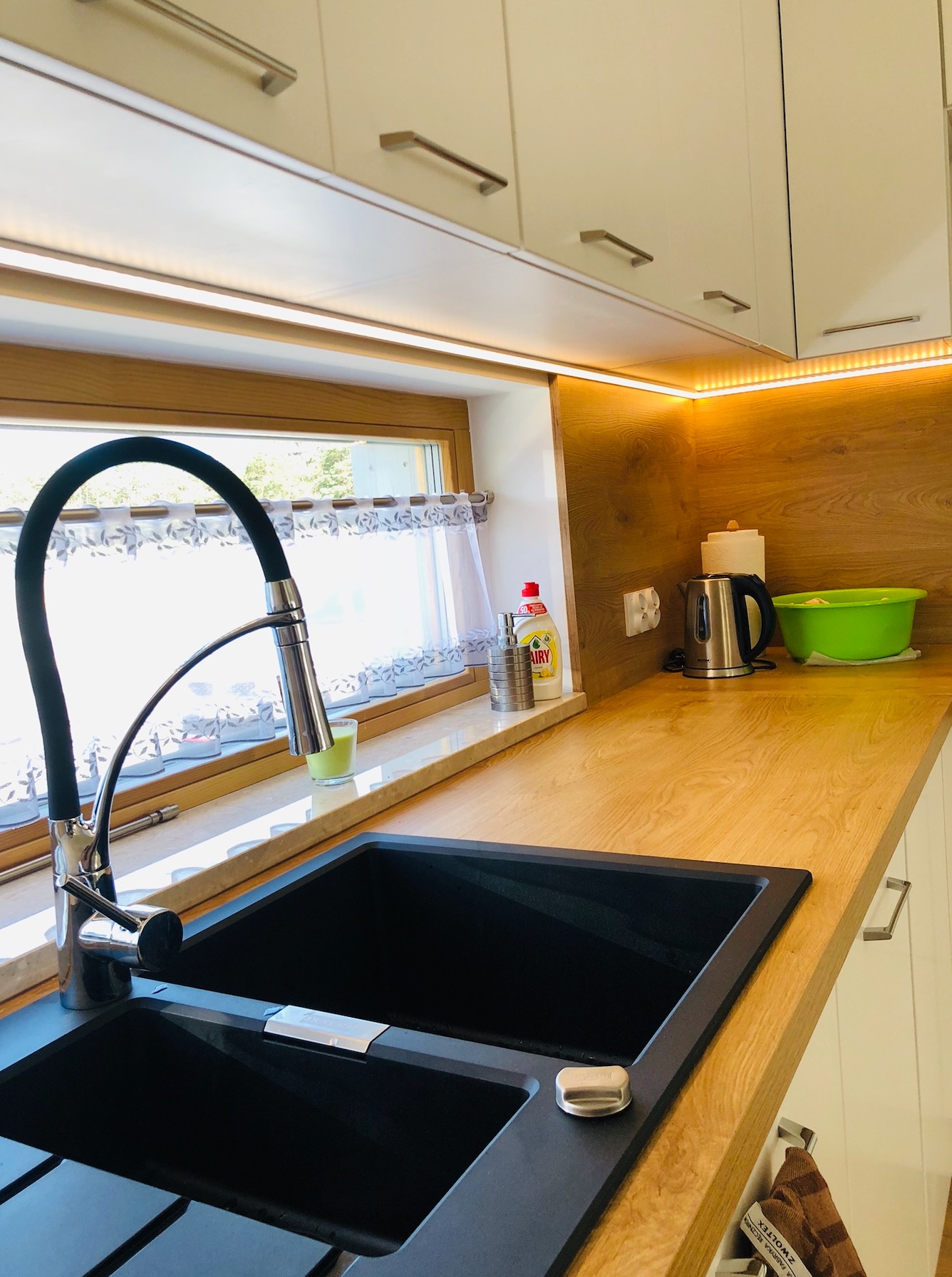 Czarny, dwukomorowy zlew z baterią w kuchni z drewnianym blatem i białymi szafkami, oświetlonymi listwami LED.