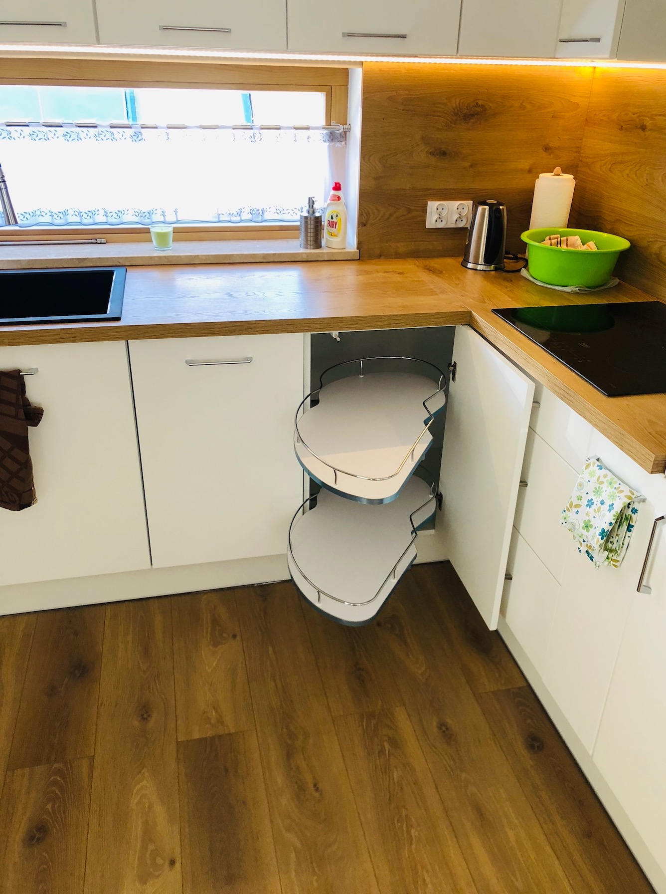 Nowoczesna kuchnia z białymi szafkami i drewnianym blatem, narożna szafka z obrotowymi półkami wysunięta, ukazująca mechanizm.