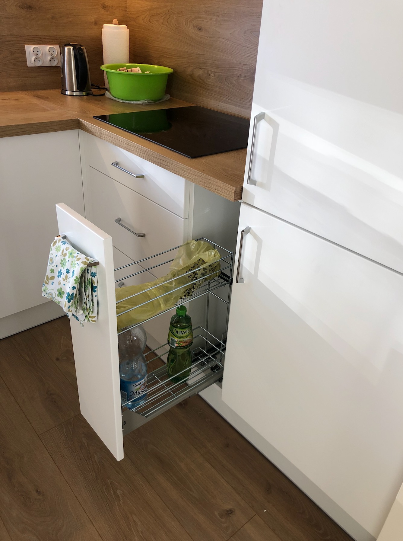 Wysuwana szafka cargo w białej zabudowie kuchennej z drewnianym blatem, widoczne butelki oleju i wody oraz ściereczka.
