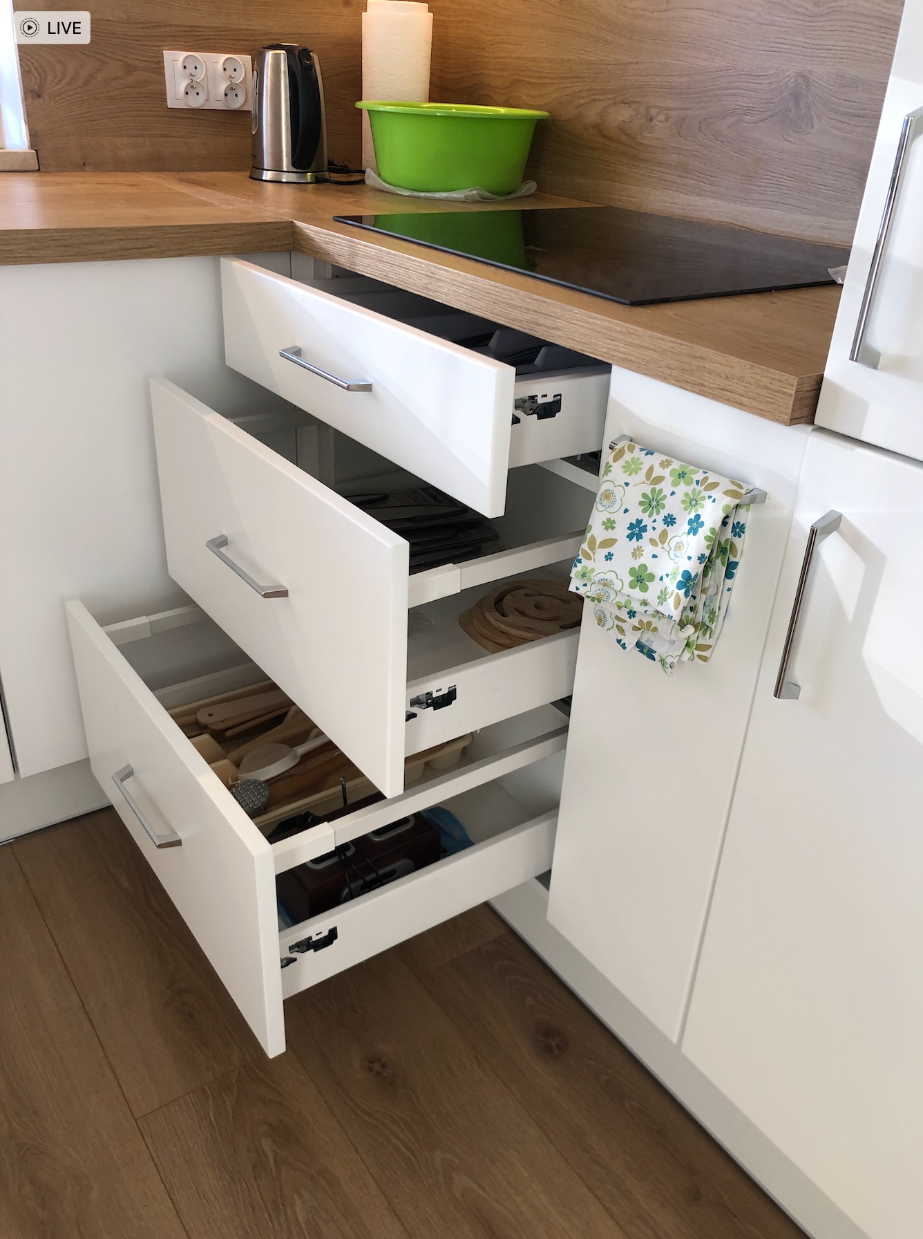 Białe, częściowo otwarte szuflady w nowoczesnej kuchni z drewnianym blatem, widoczny organizer na sztućce i akcesoria kuchenne w dolnej szufladzie, ręcznik kuchenny z kwiatowym wzorem na boku szafki.
