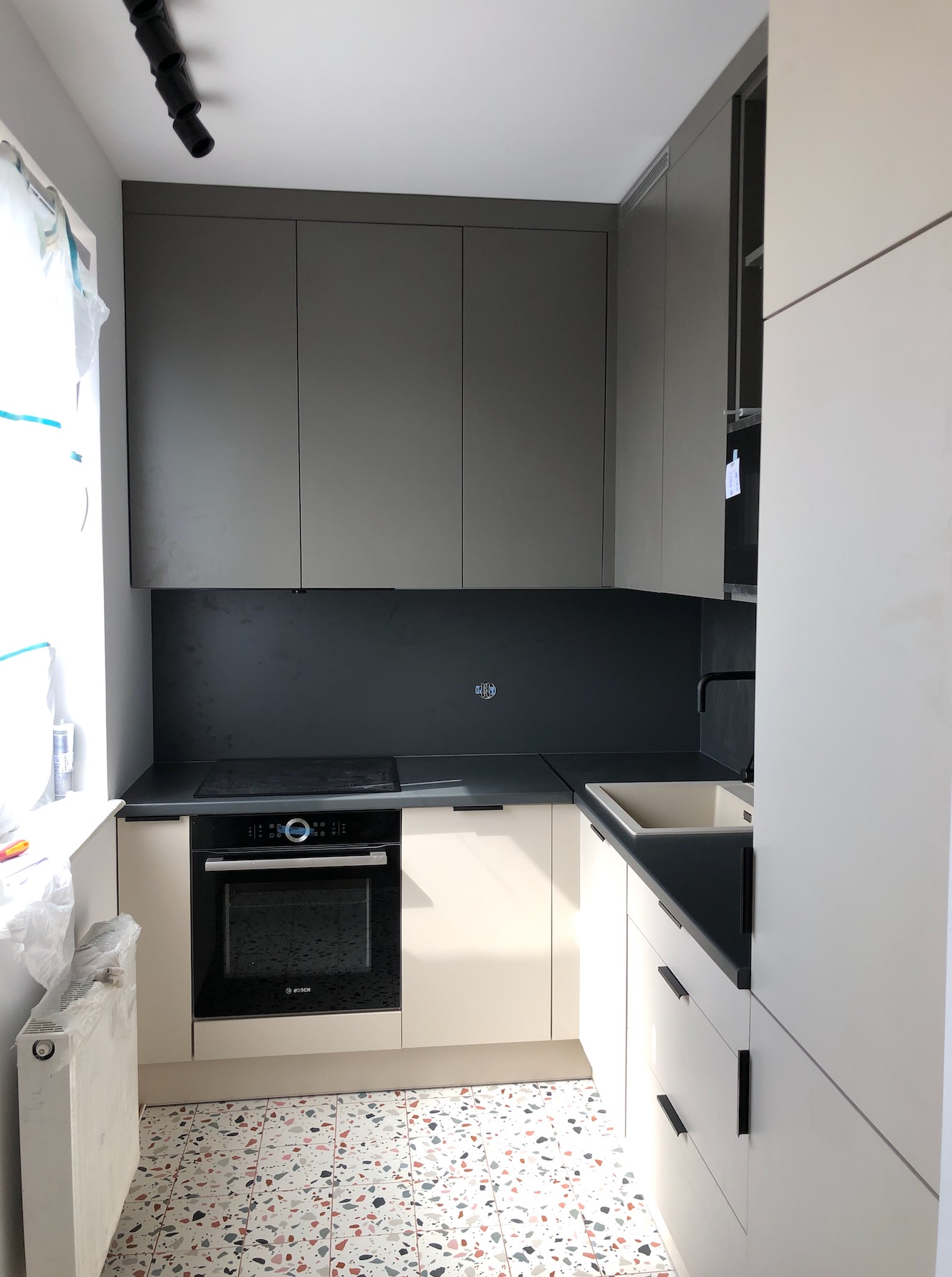 Nowoczesna, jasna kuchnia z szarymi szafkami górnymi i beżowymi dolnymi, czarnym blatem i płytą indukcyjną, widok na mozaikową podłogę i grzejnik w folii ochronnej.