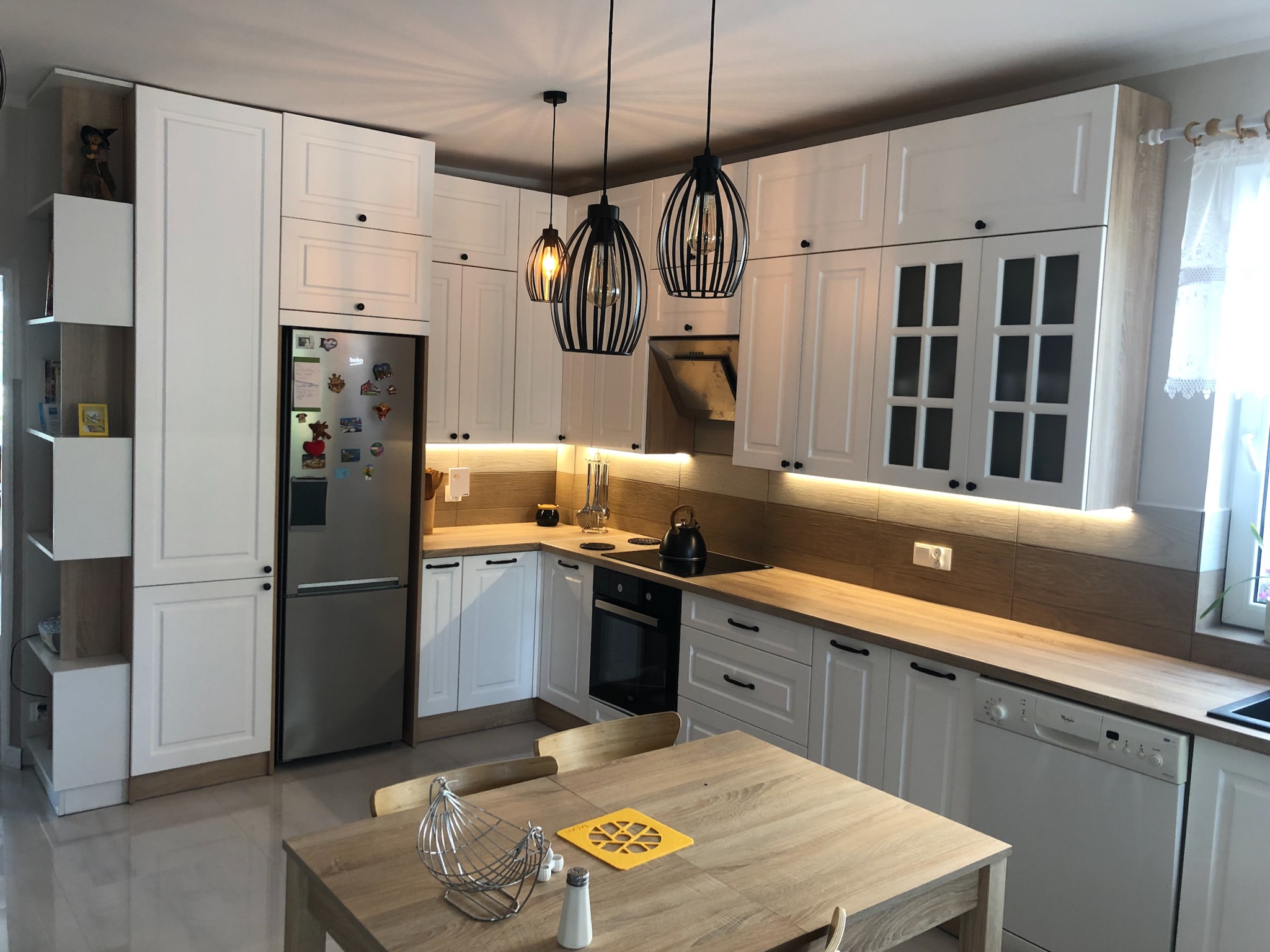 Jasna kuchnia z białymi frontami i drewnianym blatem, oświetlona trzema wiszącymi lampami w stylu industrialnym, stół z akcesoriami kuchennymi na pierwszym planie.