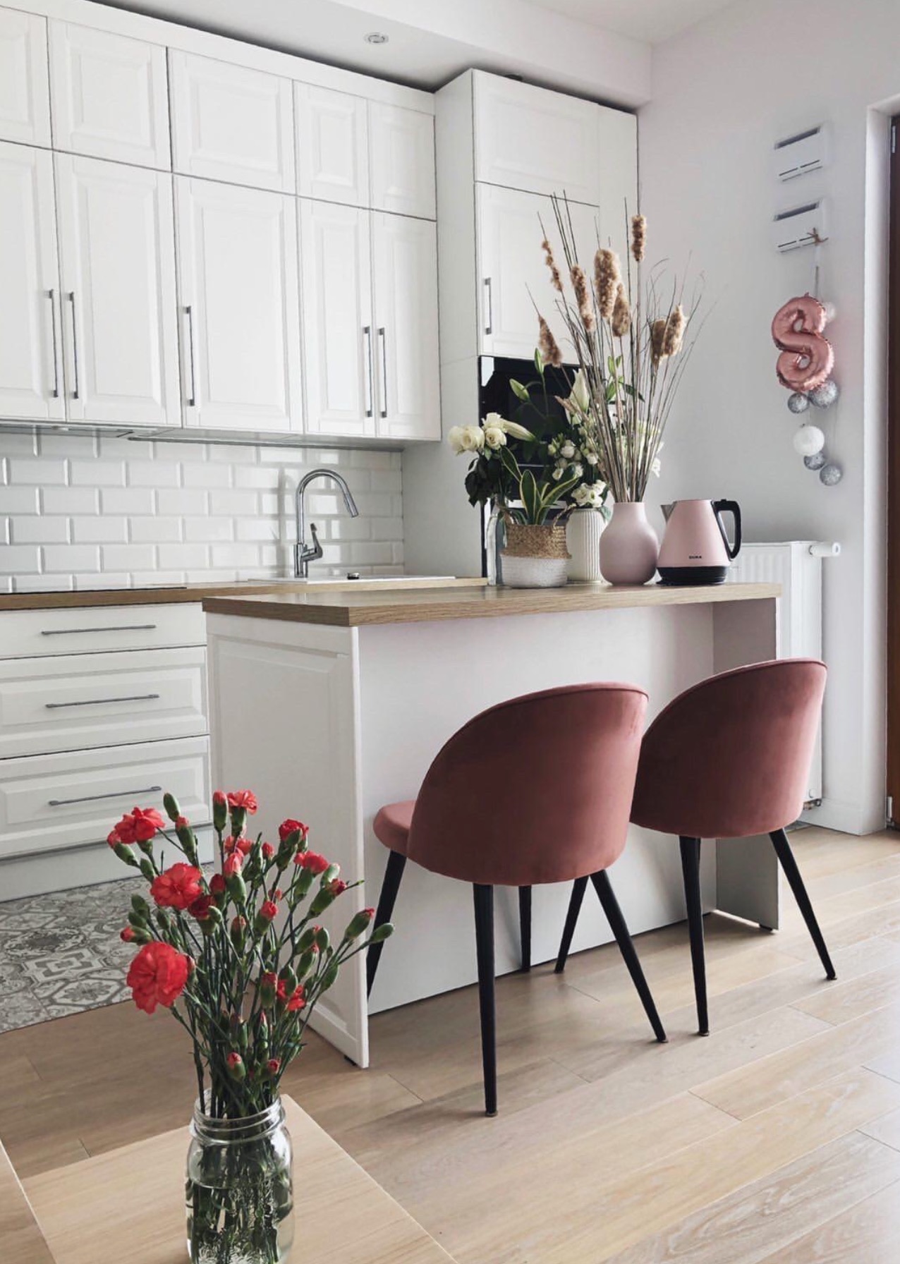 Jasna kuchnia z białymi szafkami, drewnianym blatem i dwoma różowymi krzesłami barowymi, uzupełniona dekoracjami kwiatowymi i balonem cyfrą na ścianie.