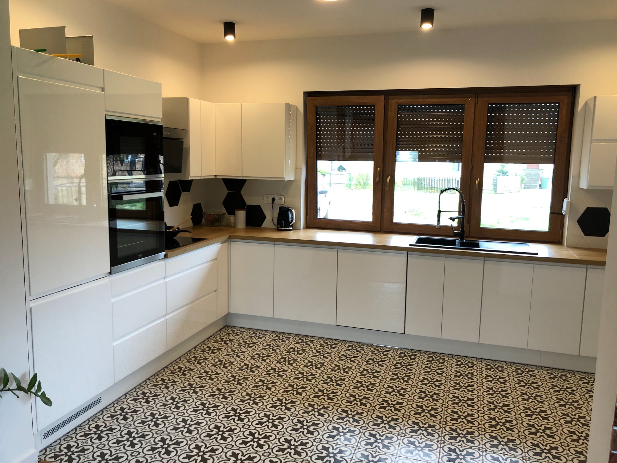 Jasna kuchnia z białymi frontami szafek, drewnianym blatem, wzorzystą podłogą i oknem z drewnianą ramą oraz roletami.