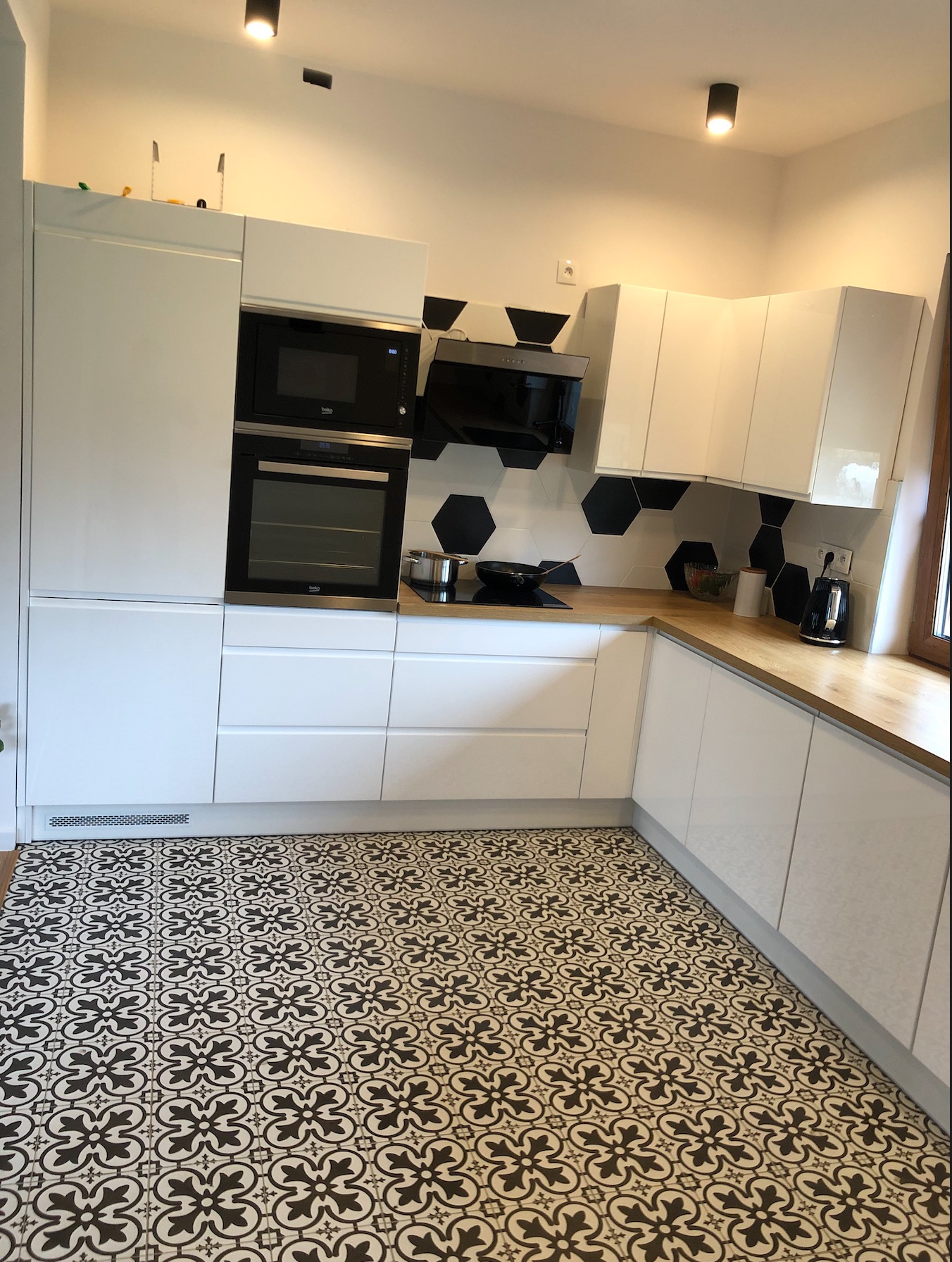 Nowoczesna kuchnia z białymi, lakierowanymi frontami, drewnianym blatem i czarno-białą, wzorzystą podłogą, uzupełniona czarnymi akcentami w postaci okapu i płytek heksagonalnych nad blatem roboczym.
