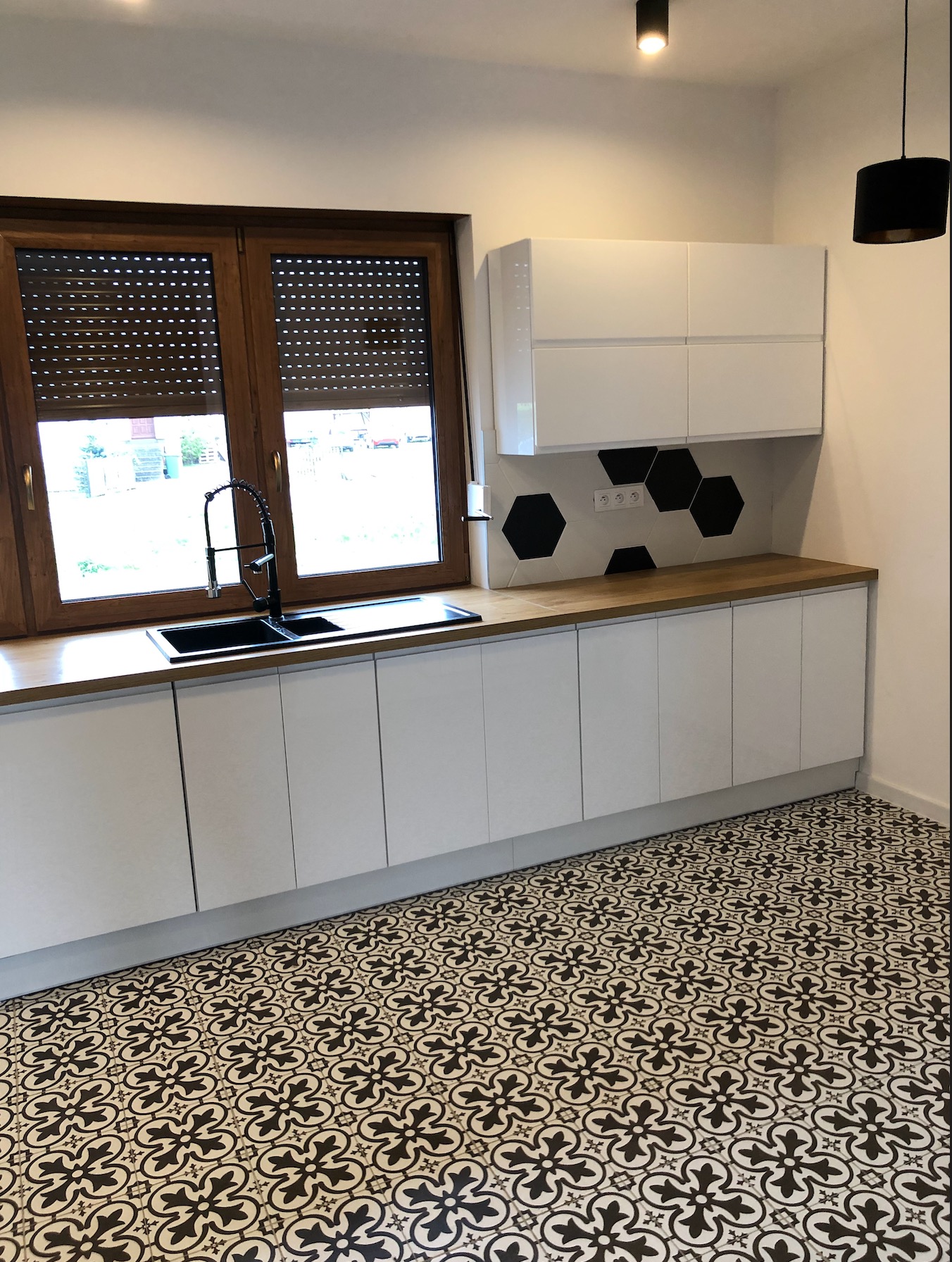 Nowoczesna kuchnia z białymi szafkami, drewnianym blatem i czarną armaturą na tle okna z brązowymi roletami, uzupełniona czarnymi heksagonalnymi płytkami i wzorzystą podłogą.