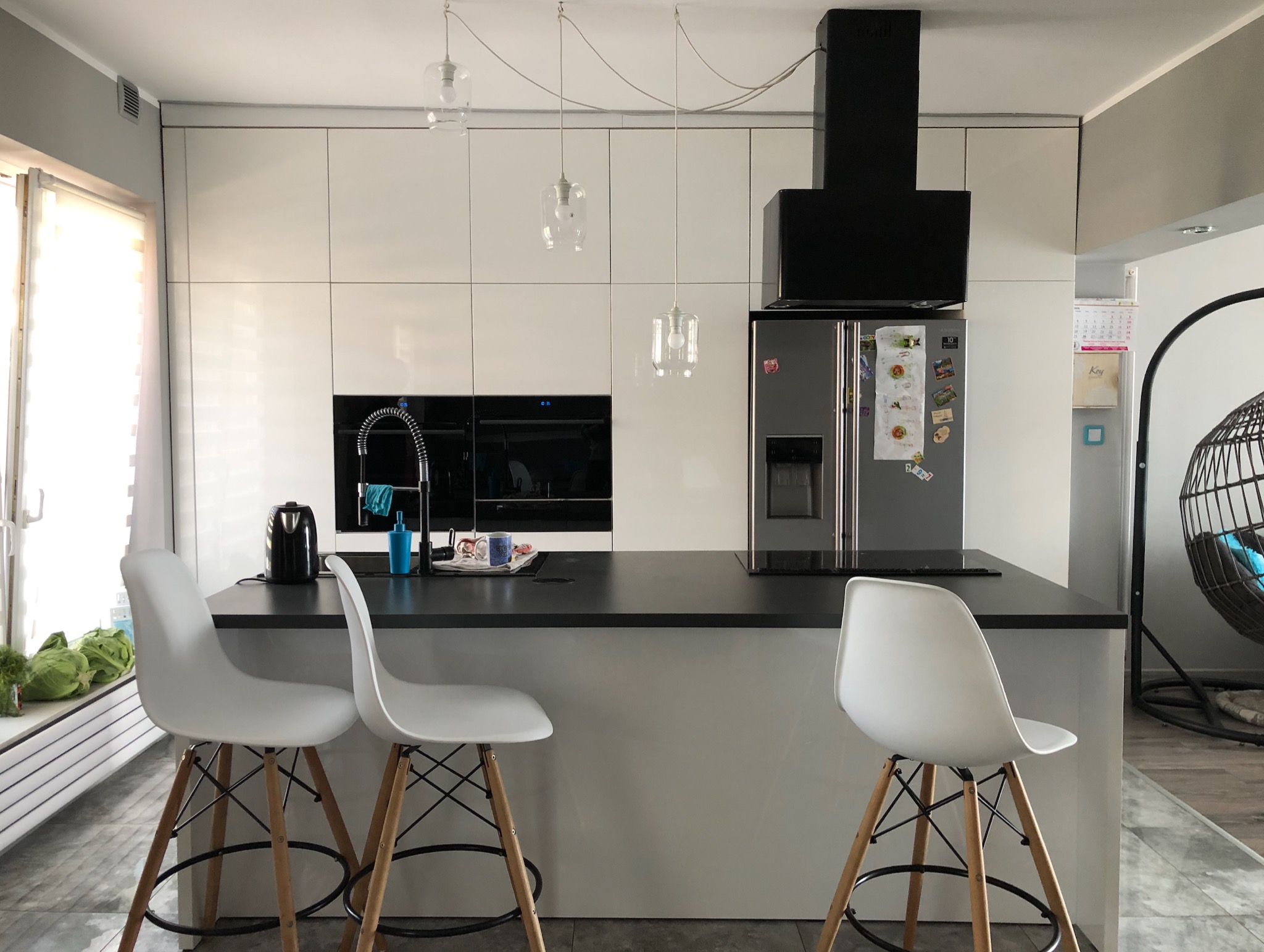 Nowoczesna kuchnia z białymi szafkami na wymiar, czarnym blatem i trzema wiszącymi lampami o przezroczystych kloszach, uzupełniona o stalową lodówkę i czarny okap.