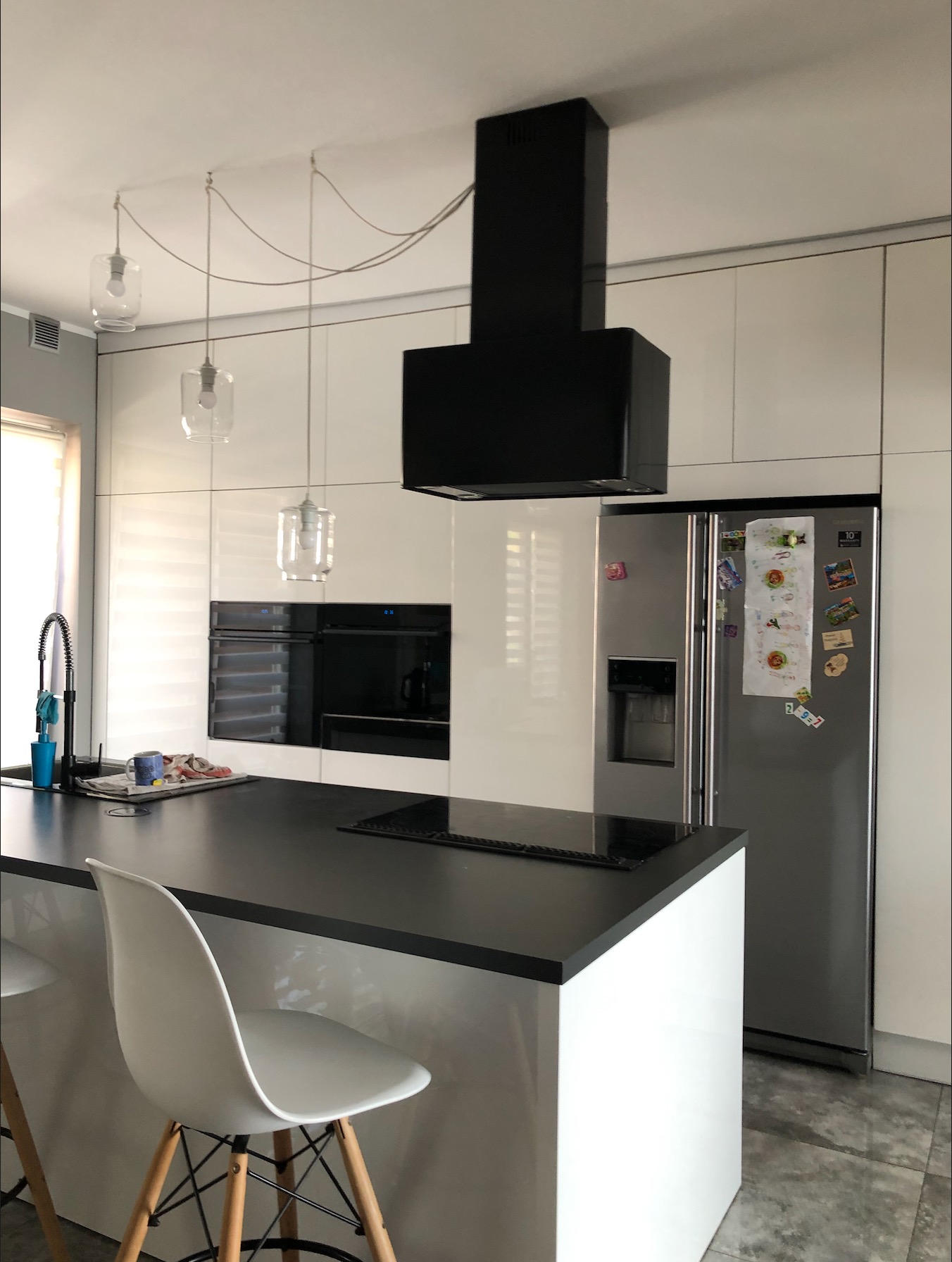 Nowoczesna kuchnia z białymi szafkami, czarnym blatem, wyspą kuchenną i czarnym okapem, oświetlona wiszącymi lampami z przezroczystego szkła na tle lodówki z magnesami.