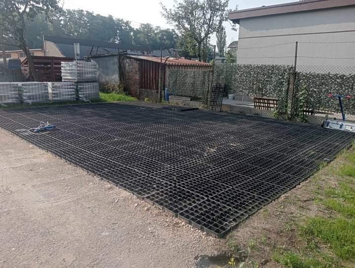 Ułożona, czarna, plastikowa kratka trawnikowa na przygotowanym podłożu z piasku, widoczne palety z kostką brukową w tle oraz budynki gospodarcze.