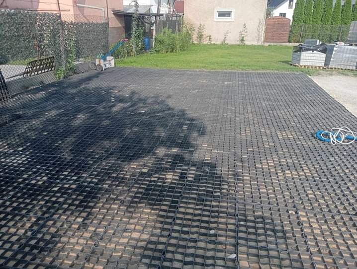 Ułożona, czarna, ażurowa nawierzchnia na piasku, widoczne cienie drzew, w tle trawnik i budynek.