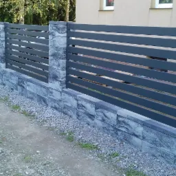 Nowoczesne ogrodzenie z poziomych, ciemnoszarych paneli metalowych, osadzone na niskim murku z szarych, wzorzystych bloczków betonowych, z widocznym podłożem z drobnego żwiru.