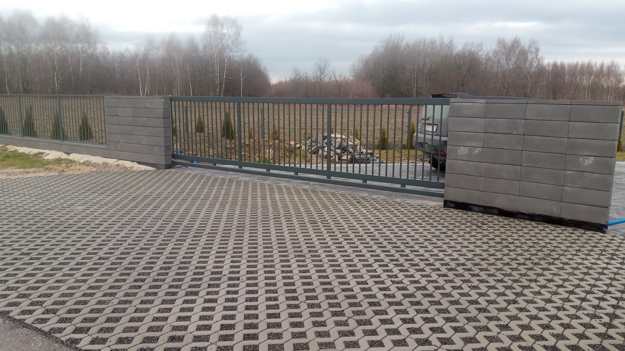 Nowoczesna brama wjazdowa przesuwna, stalowa, szara, z wypełnieniem z pionowych prętów, zamontowana między słupkami z szarej kostki betonowej, widok na podjazd z ażurowych płyt betonowych i drzewa...