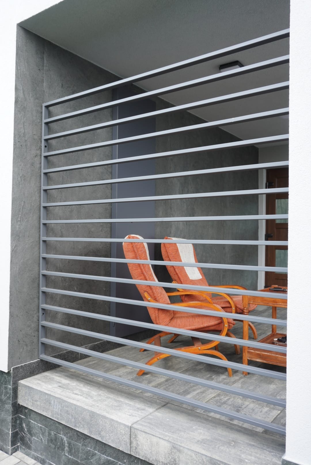Szary, poziomy układ prętów tworzących osłonę tarasu, za którą widoczne są dwa fotele z pomarańczową tapicerką i drewniany stolik.