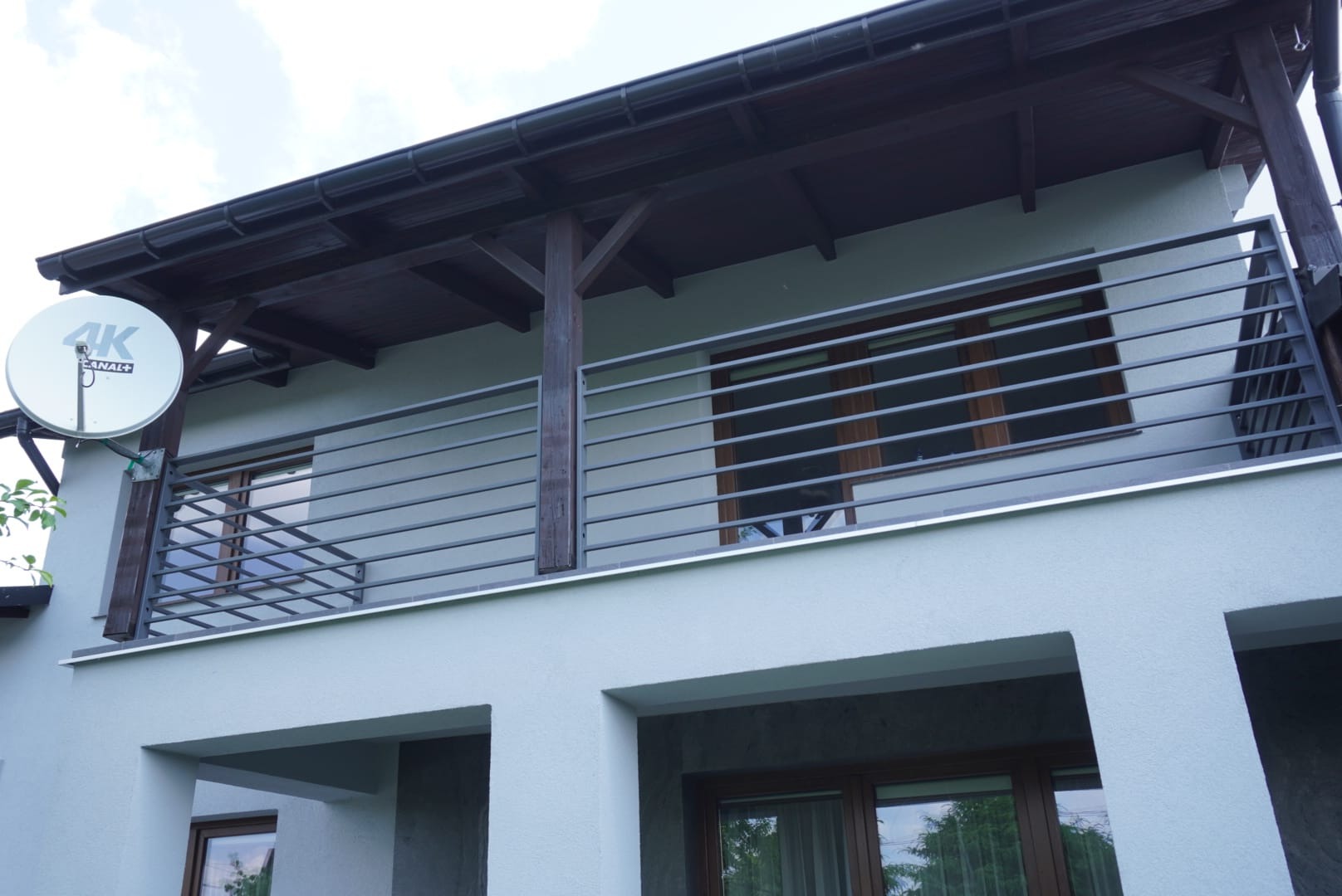 Szara elewacja domu z balkonem zabezpieczonym poziomą, metalową balustradą w kolorze grafitowym, z widoczną anteną satelitarną na ścianie.